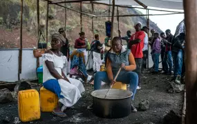 Two women sit around a large pot. One of them stirs the contents of it with a wooden stick. In the back-drop we see individuals standing by.